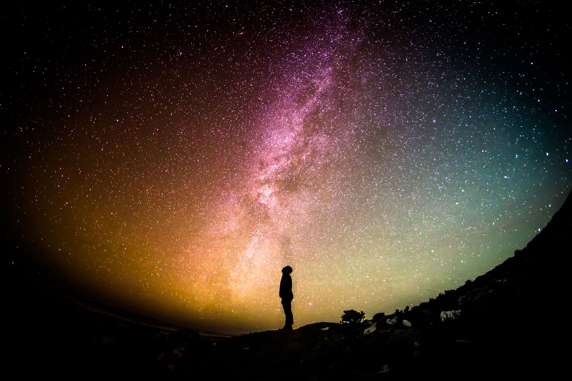Ein Mann steht im Profil  in der Nacht vor einem kugelförmigen hell erleuchteten, bunten Sternenhimmel und schaut nach oben. Ein Symbol der unendlichen Möglichkeiten, die du dir aussuchen kannst, wer du sein möchtest. Hierbei geht es um den Glauben, wer du bist. 